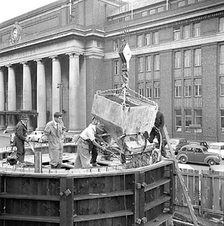 d. views of work in progress, workmen pouring concrete using an overhead crane. Railway Station in background