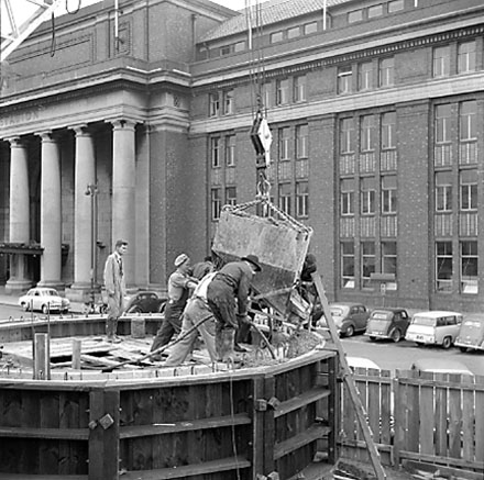 e. views of work in progress, workmen pouring concrete using an overhead crane. Railway Station in background