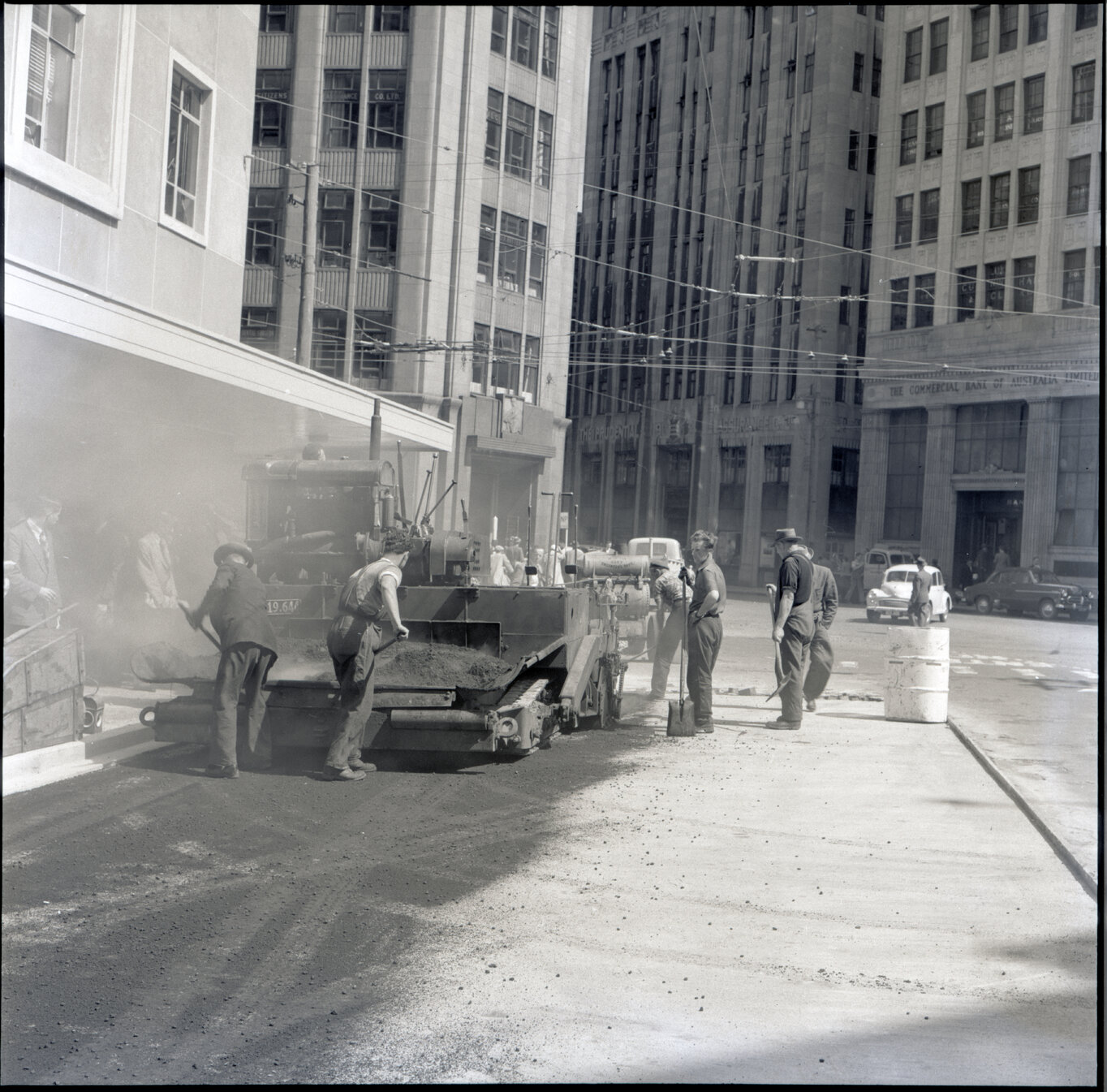 f. Various stages of asphaltic road surfacing by Council workmen, Lambton Quay end of Featherston Street. People passing by have stopped to watch work in progress. Trolly buses in background