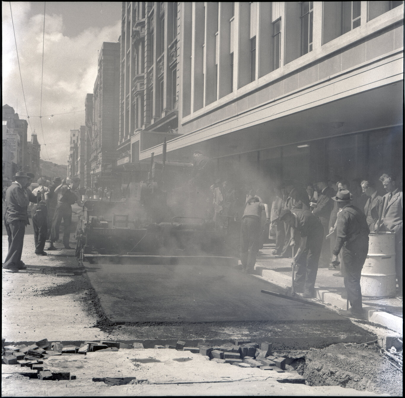 e. Various stages of asphaltic road surfacing by Council workmen, Lambton Quay end of Featherston Street. People passing by have stopped to watch work in progress. Trolly buses in background