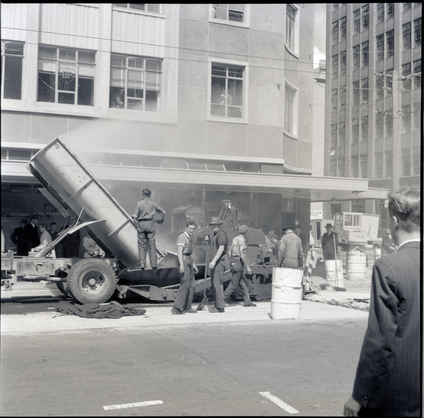 g. Various stages of asphaltic road surfacing by Council workmen, Lambton Quay end of Featherston Street. People passing by have stopped to watch work in progress. Trolly buses in background
