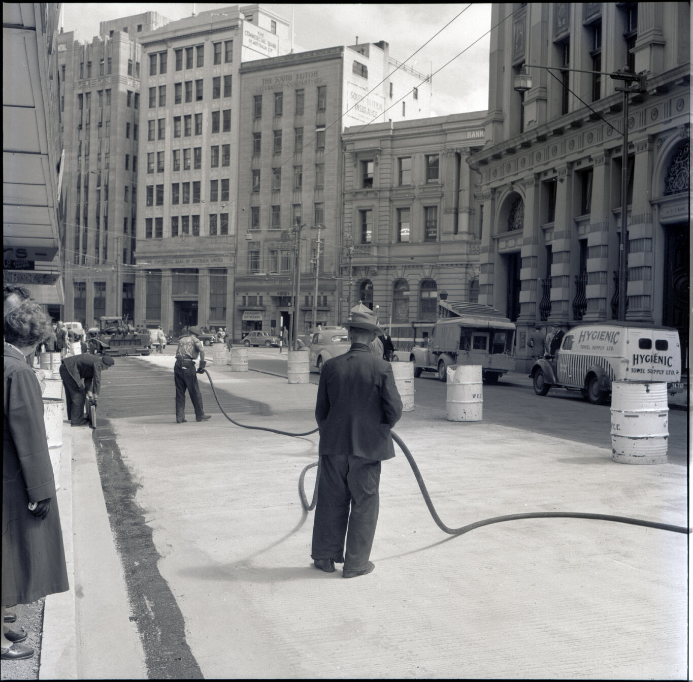i. Various stages of asphaltic road surfacing by Council workmen, Lambton Quay end of Featherston Street. People passing by have stopped to watch work in progress. Trolly buses in background