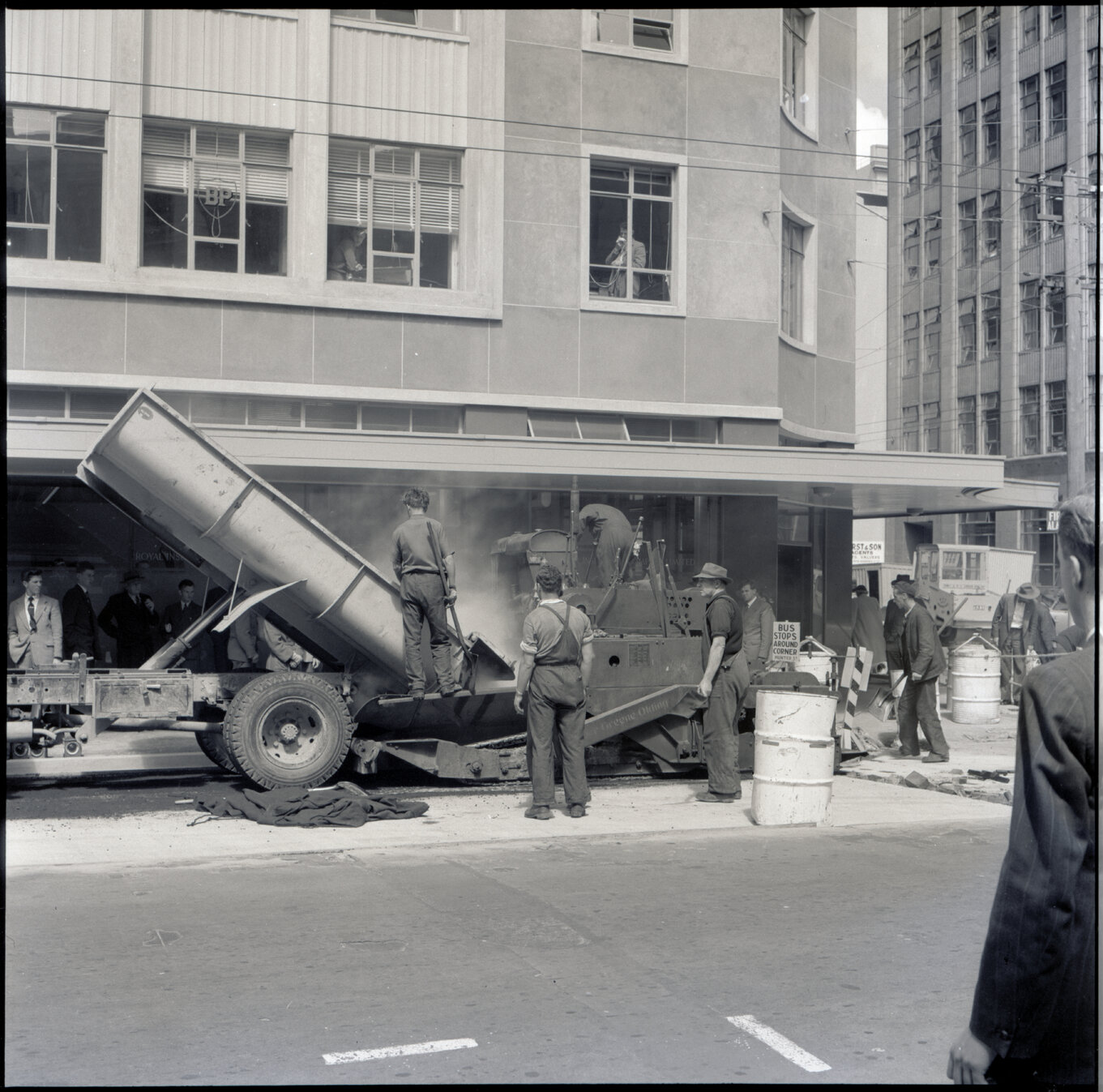h. Various stages of asphaltic road surfacing by Council workmen, Lambton Quay end of Featherston Street. People passing by have stopped to watch work in progress. Trolly buses in background