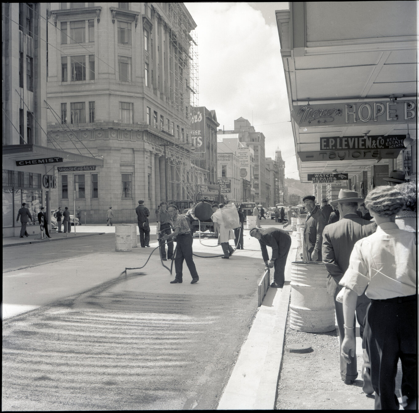 j. Various stages of asphaltic road surfacing by Council workmen, Lambton Quay end of Featherston Street. People passing by have stopped to watch work in progress. Trolly buses in background