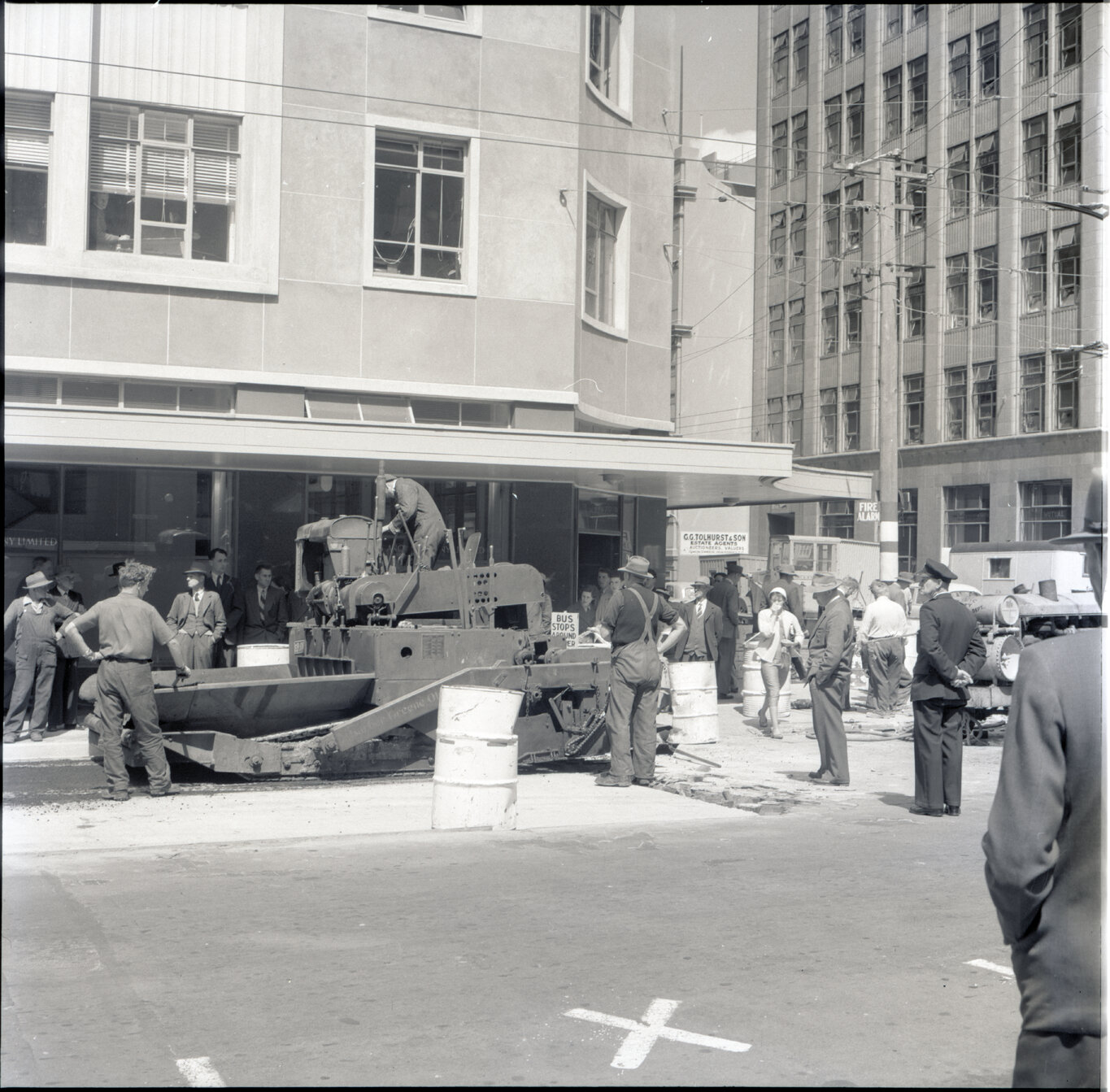 k. Various stages of asphaltic road surfacing by Council workmen, Lambton Quay end of Featherston Street. People passing by have stopped to watch work in progress. Trolly buses in background