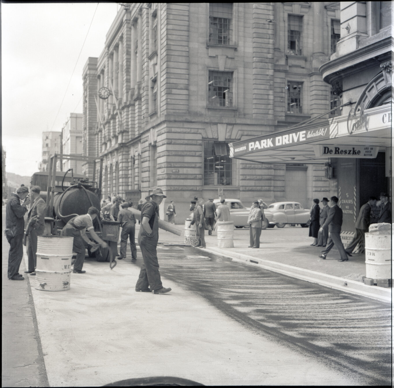 l. Various stages of asphaltic road surfacing by Council workmen, Lambton Quay end of Featherston Street. People passing by have stopped to watch work in progress. Trolly buses in background