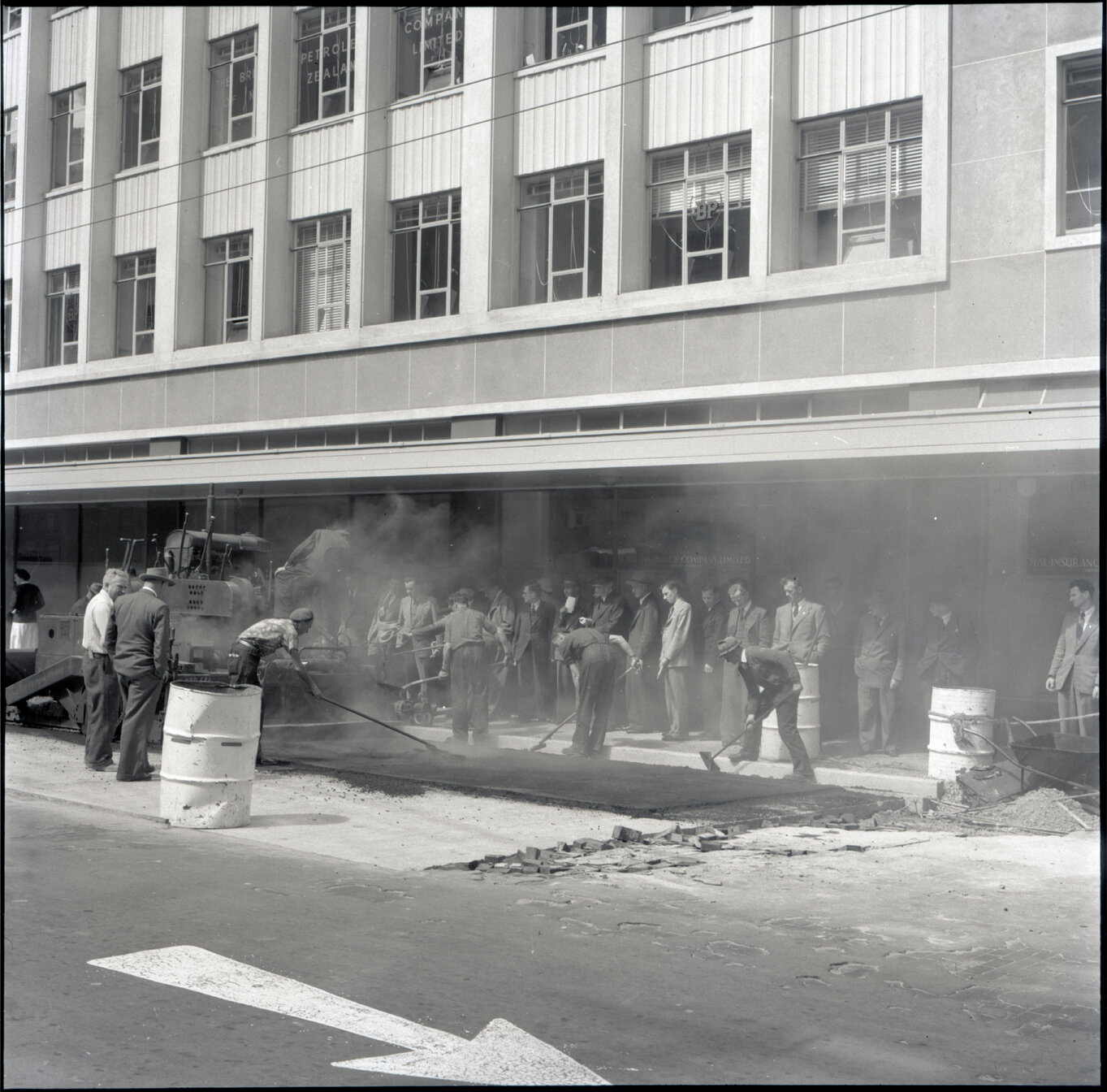 n. Various stages of asphaltic road surfacing by Council workmen, Lambton Quay end of Featherston Street. People passing by have stopped to watch work in progress. Trolly buses in background