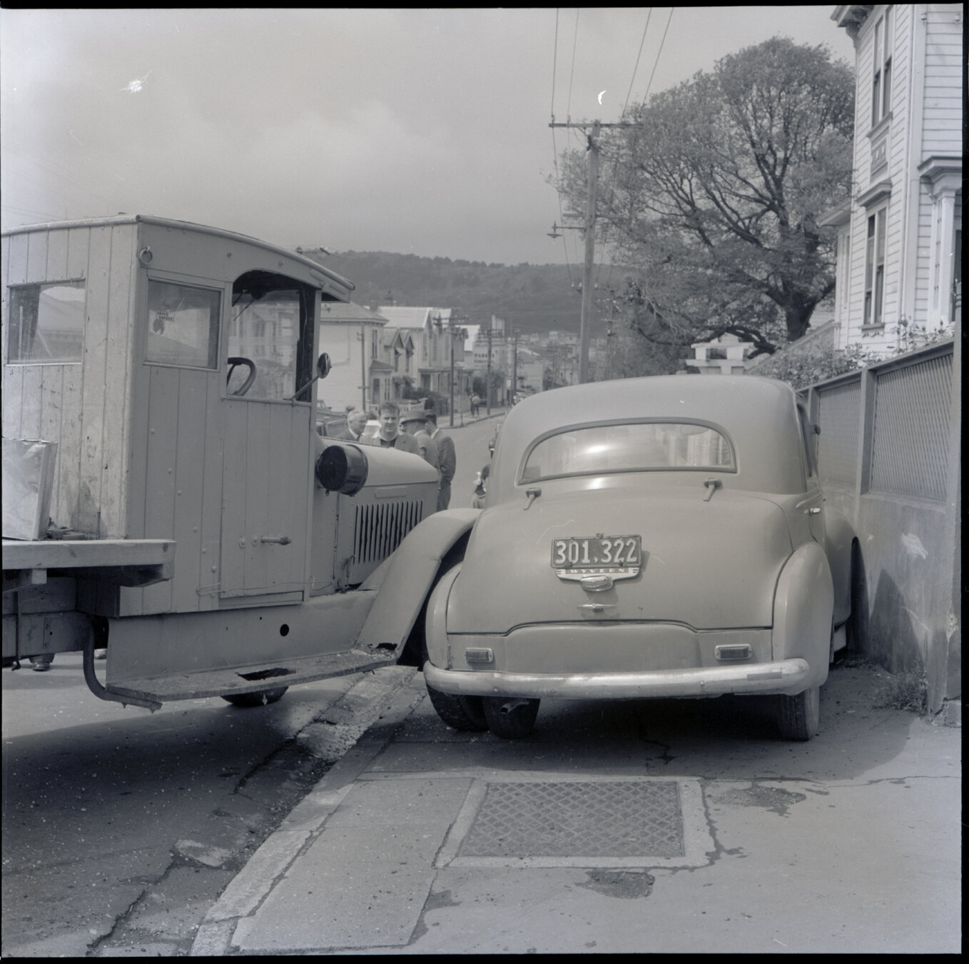 d. A flatbed truck and motor vehicle have collided, at the corner of Willis Street and Abel Smith Street. People, including men in suits and a group of young boys, have gathered. 319 Willis Street, a two storey wooden building is in the background. Gilby'