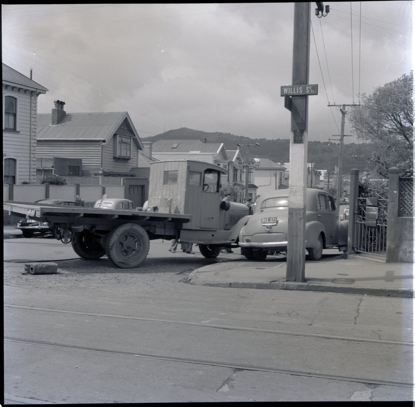 f. A flatbed truck and motor vehicle have collided, at the corner of Willis Street and Abel Smith Street. People, including men in suits and a group of young boys, have gathered. 319 Willis Street, a two storey wooden building is in the background. Gilby'