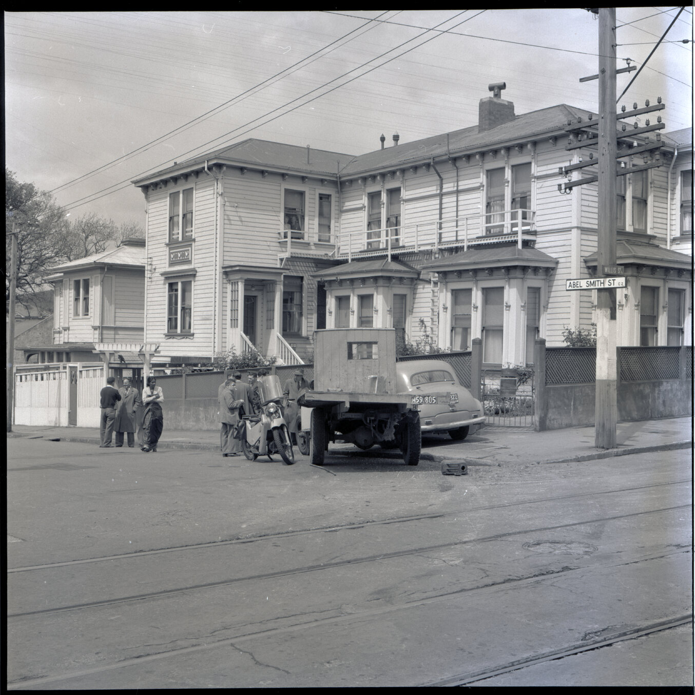 g. A flatbed truck and motor vehicle have collided, at the corner of Willis Street and Abel Smith Street. People, including men in suits and a group of young boys, have gathered. 319 Willis Street, a two storey wooden building is in the background. Gilby'