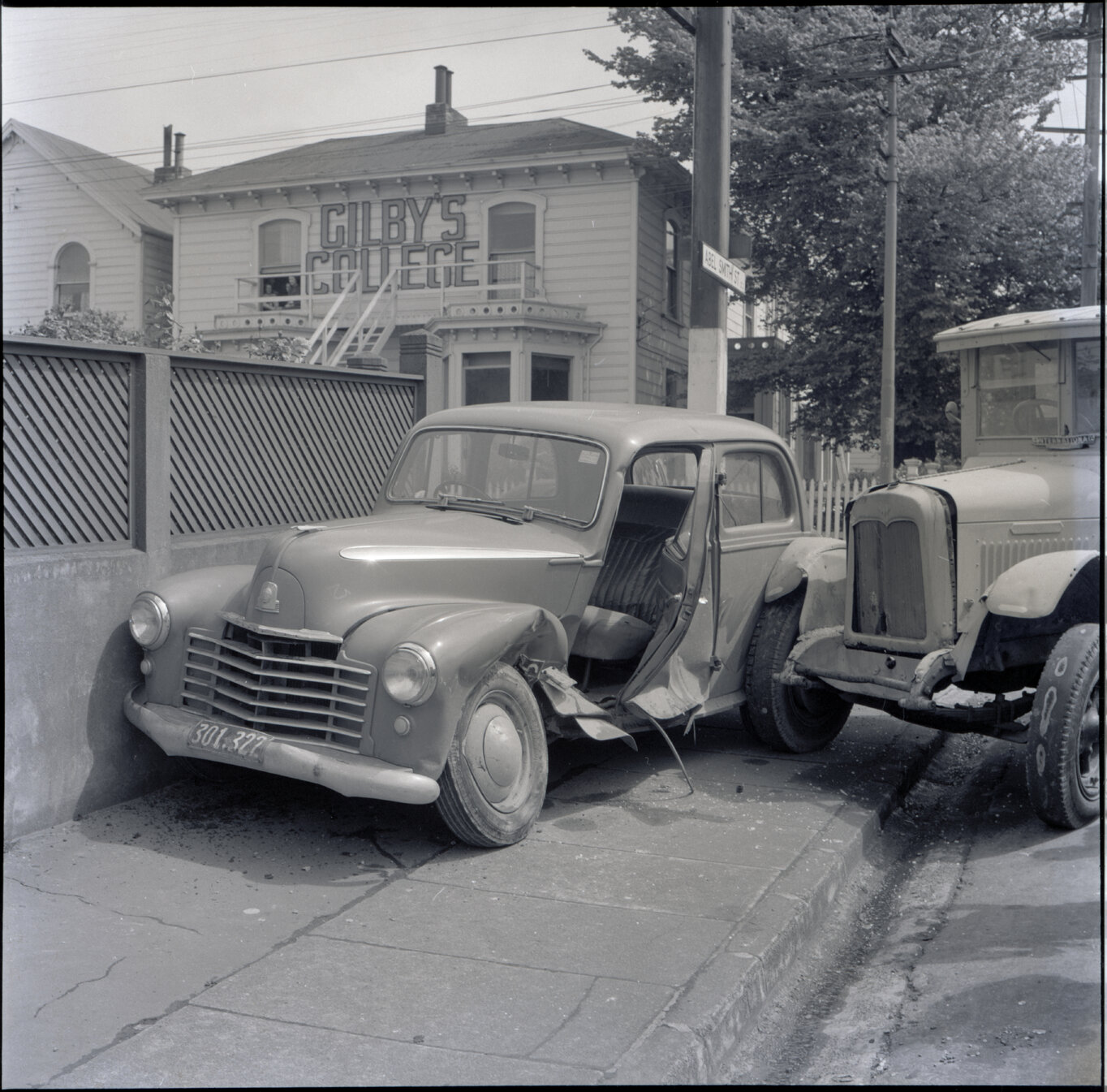 a. A flatbed truck and motor vehicle have collided, at the corner of Willis Street and Abel Smith Street. People, including men in suits and a group of young boys, have gathered. 319 Willis Street, a two storey wooden building is in the background. Gilby'