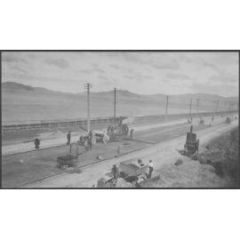 Road surface is being prepared for resurfacing, steam roller is in the background.